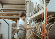 A woman in a white t-shirt and gray pants stands in a workshop or factory next to a large machine, holding a tablet and reaching toward shelves filled with rolls of material.
