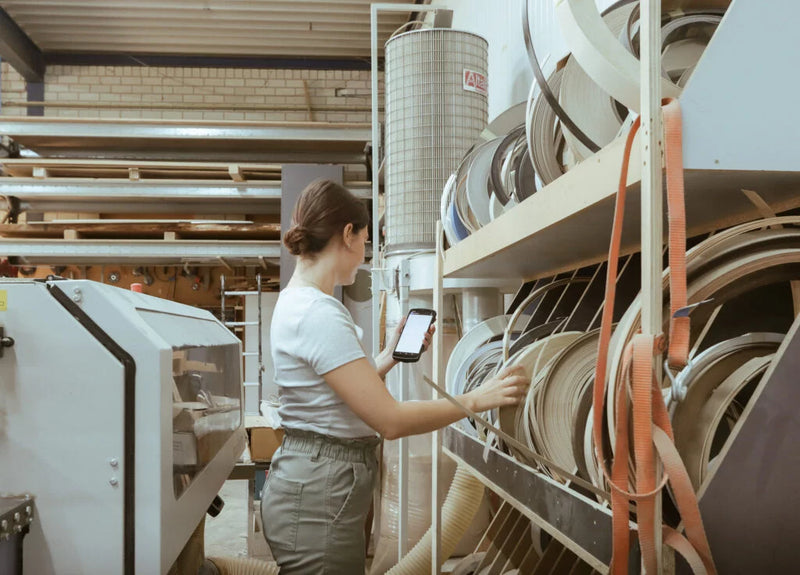 A woman in a white t-shirt and gray pants stands in a workshop or factory next to a large machine, holding a tablet and reaching toward shelves filled with rolls of material.