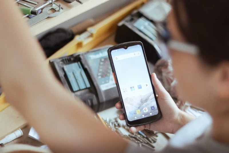 A person in a workshop holding a black smartphone with a lit screen, with tools and equipment blurred in the background.